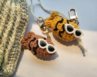 May include: Two crocheted keychains, one brown and one yellow, each with white sunglasses and black detailing. The keychains are attached to gold-colored clasps. A crocheted bag is visible in the background.
