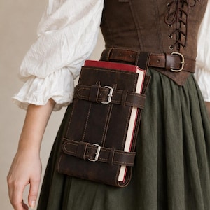 May include: A dark brown leather book holder with two silver-toned buckles, attached to a matching brown leather belt. The holder secures a book with a red spine. The ensemble is worn over a dark brown corset with front lacing and a long green skirt.