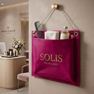 May include: A magenta wall-mounted storage pocket featuring the gold text SOLIS YOUR LOGO. The pocket hangs from a gold circular hook by a thin cord and contains a black comb, a white folded cloth, and a clear bottle labelled Eclat Rose.