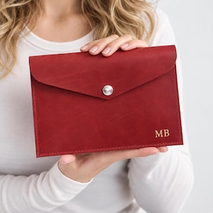 May include: A deep red leather envelope-style clutch featuring a pointed flap with a single silver-toned snap button closure. The bottom right corner of the front panel is embossed with the gold-colored capital letters MB in a serif font.