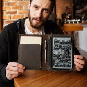 Könnte beinhalten: Eine dunkelbraune Leder-E-Reader-Hülle mit eingebautem Notizblock und Stift. Der E-Reader zeigt einen Schwarz-Weiß-Comic. Die Hülle ist geöffnet und gibt den Inhalt frei. Die Hülle liegt auf einem Holztisch.