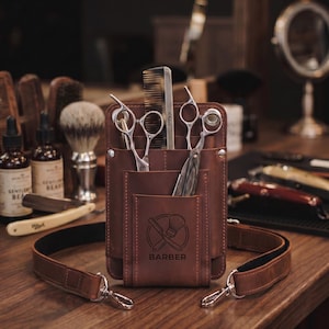 May include: A brown leather barber tool holster with an adjustable shoulder strap. The front features a stamped logo of a straight razor and shaving brush with the text BARBER. It holds two pairs of silver metal scissors and a black comb.