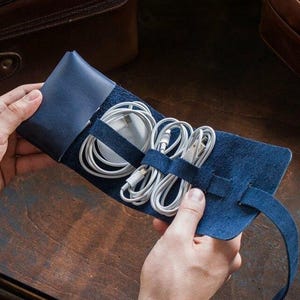 May include: A blue leather cable organizer holding white cables. The organizer is open, revealing the neatly arranged cords. The background includes brown leather bags and a wooden surface.