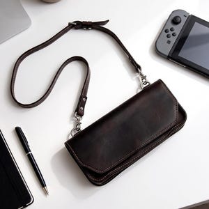 May include: A dark brown leather crossbody bag with a rectangular flap, visible stitching, and a thin adjustable shoulder strap attached with silver-toned metal hardware clips. The bag features a smooth finish and a structured, elongated rectangular shape.