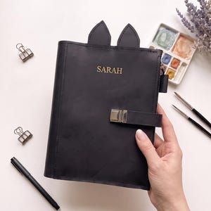 Puede incluir: Una funda de cuaderno de cuero negro con dos orejas puntiagudas de gato en el borde superior. La parte frontal tiene grabado el nombre SARAH en letras doradas e incluye una correa horizontal con un cierre de hebilla de metal plateado.