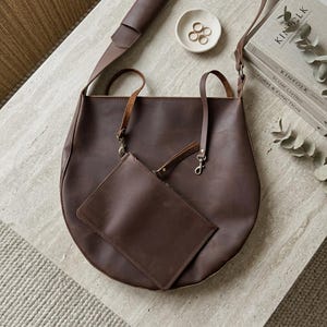 May include: A dark brown leather tote bag with a matching detachable zippered pouch. The bag features thin shoulder straps, metal swivel clasps, and a smooth matte finish. The design includes a long adjustable shoulder strap attached to the top.
