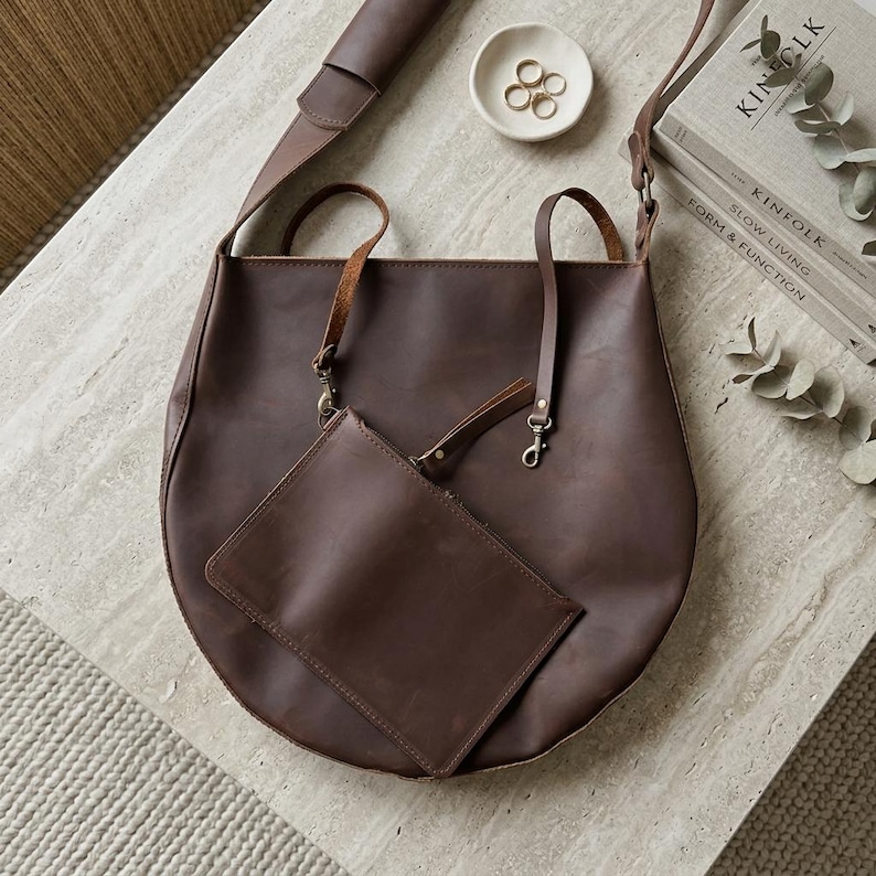 May include: A dark brown leather tote bag with a matching detachable zippered pouch. The bag features thin shoulder straps, metal swivel clasps, and a smooth matte finish. The design includes a long adjustable shoulder strap attached to the top.
