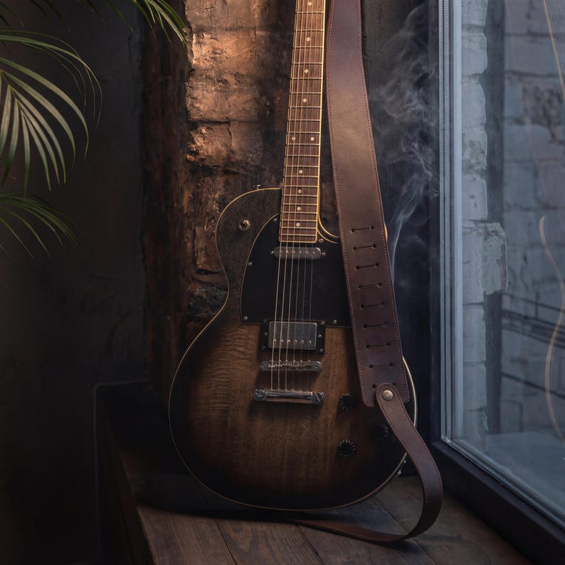 K&ouml;nnte beinhalten: Eine E-Gitarre mit dunkelbraunem und schwarzem Sunburst-Finish, ausgestattet mit einem schwarzen Schlagbrett, silberner Hardware und sechs Saiten. Ein breiter, dunkelbrauner Ledergitarrengurt mit vertikalen Einstellschlitzen ist am Korpus befestigt.
