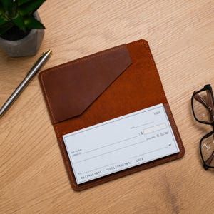 Op de afbeelding: Een bruine leren chequeboekhoes met een diagonaal vakje, met daarin een witte papieren cheque met het nummer 1001 in de rechterbovenhoek. De cheque bevat standaardvelden voor datum, begunstigde, bedrag en handtekening.