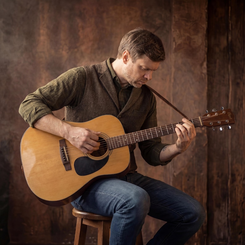 K&ouml;nnte beinhalten: Eine helle Akustikgitarre mit einem dunklen Schlagbrett, sechs Stahlsaiten und einem Holzgriffbrett. Das Instrument verf&uuml;gt &uuml;ber ein nat&uuml;rliches Holzfinish, ein rundes Schallloch und silberne Stimmwirbel an der Kopfplatte der Gitarre.