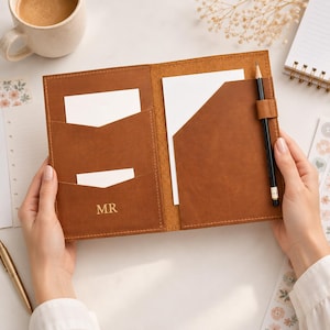 Puede incluir: Una funda de cuero color canela para cuaderno con dos bolsillos interiores, uno de ellos grabado con las letras MR. La funda contiene hojas de papel blanco e incluye un lápiz negro sujeto en una trabilla de cuero en el lado derecho.