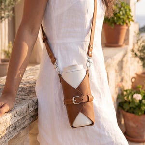 May include: A tan leather bottle holder with a matching adjustable strap and a silver-toned metal buckle closure. The holder features a wrap-around design that secures a white cylindrical bottle, suspended by silver-toned swivel clips on each side.