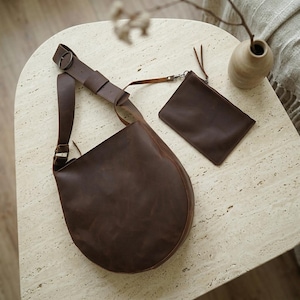 May include: A dark brown leather hobo bag with a wide adjustable shoulder strap featuring a metal buckle and a matching rectangular leather pouch attached by a thin strap. The bag has a smooth, matte finish and a rounded, crescent-like shape.