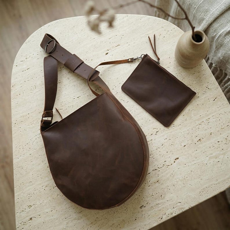May include: A dark brown leather hobo bag with a wide adjustable shoulder strap featuring a metal buckle and a matching rectangular leather pouch attached by a thin strap. The bag has a smooth, matte finish and a rounded, crescent-like shape.