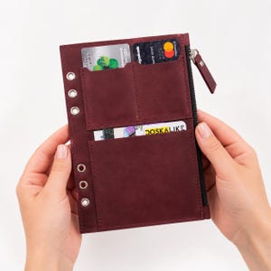 May include: A deep burgundy leather planner insert featuring a vertical row of six silver metal binder rings on the left side, two top card slots, one large bottom pocket, and a black zippered compartment on the right side for secure storage.