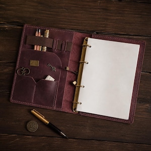 Puede incluir: Una funda de diario de cuero morado oscuro con un mecanismo de anillas de latón que sujeta páginas blancas en blanco. El interior cuenta con varios bolsillos, trabillas para bolígrafos con dos lápices y un bolígrafo dorado, y pequeños dijes.