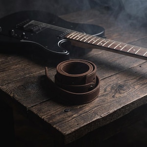 K&ouml;nnte beinhalten: Ein aufgerollter Gitarrengurt aus dunkelbraunem Leder mit sichtbaren N&auml;hten und einem metallischen Knopfverschluss, platziert neben einer schwarzen E-Gitarre auf einer dunklen Holzoberfl&auml;che mit einem dezenten Raucheffekt im Hintergrund.
