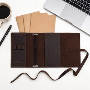 Puede incluir: Una funda de cuero marrón oscuro para cuaderno de viaje con el nombre LUKE grabado en el panel frontal izquierdo. El interior incluye tres bandas elásticas para insertos, un portalápices y varias ranuras para tarjetas, con cierre de tira de cuero.