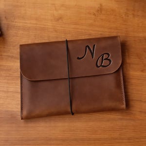 Op de afbeelding: Een bruine leren portfolio met een klepsluiting en een zwarte elastische band. De initialen "N B" zijn op de voorkant in reliëf aangebracht. Ontworpen voor documenten of een tablet.