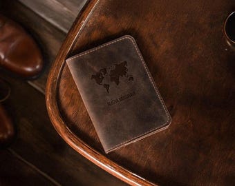 May include: A brown leather passport holder with a world map design and the name "ALISHA MUSGRAVE" embossed on the front. The passport holder is on a wooden table, with a pair of brown leather shoes in the background.