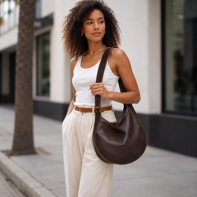 May include: A dark brown leather hobo bag with a wide shoulder strap and gold-toned hardware. The bag features a curved, crescent-shaped silhouette and a smooth, matte finish, worn over the shoulder with a white tank top and beige trousers.