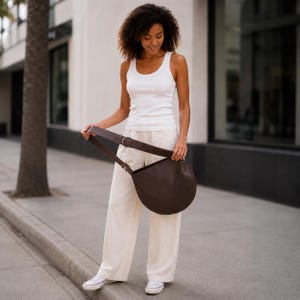 May include: A dark brown crescent-shaped shoulder bag with a wide strap and a circular metal buckle, worn with a white sleeveless tank top, loose-fitting off-white trousers, and white low-top canvas sneakers with white laces and rubber soles.