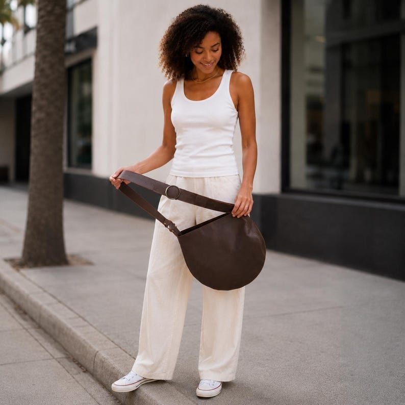 May include: A dark brown crescent-shaped shoulder bag with a wide strap and a circular metal buckle, worn with a white sleeveless tank top, loose-fitting off-white trousers, and white low-top canvas sneakers with white laces and rubber soles.