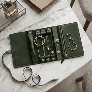 K&ouml;nnte beinhalten: Ein dunkelgr&uuml;ner Schmuck-Organizer aus Leder mit mehreren Riemen mit Druckkn&ouml;pfen f&uuml;r Halsketten und Ringe, einer Rei&szlig;verschlusstasche und speziellen Ohrringl&ouml;chern. Das flache, rechteckige Etui verf&uuml;gt &uuml;ber ein langes Lederband zum Verschlie&szlig;en.
