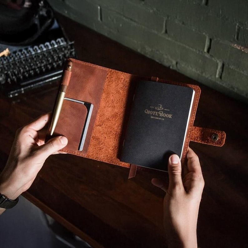 Puede incluir: Funda de cuaderno de cuero marr&oacute;n abierta con un cuaderno negro en el interior. Se ve un bol&iacute;grafo dorado y un peque&ntilde;o tarjetero. El cuaderno negro tiene las palabras "QUOTE BOOK" impresas en dorado. La funda se asegura con un cierre a presi&oacute;n.