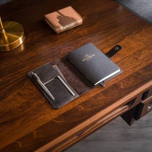 Puede incluir: Un juego de cuaderno y cartera de cuero marr&oacute;n oscuro sobre un escritorio de madera. El cuaderno est&aacute; cerrado con una correa y la cartera est&aacute; abierta, revelando un bol&iacute;grafo y tarjetas. Tambi&eacute;n es visible un art&iacute;culo de cuero m&aacute;s peque&ntilde;o de color marr&oacute;n claro.