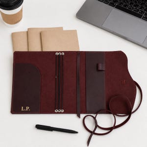 May include: A dark burgundy leather journal cover featuring three internal elastic bands, a vertical leather bookmark, a pen loop, and a side pocket embossed with the gold initials L.P. The cover includes a long leather wrap-around tie.