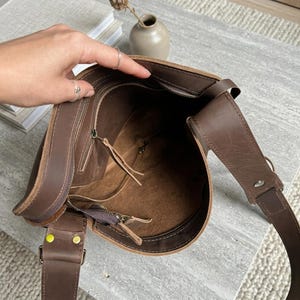 May include: A dark brown leather bucket bag with an open top, featuring a smooth interior lining, a zippered side pocket with a leather pull tab, and a wide adjustable shoulder strap attached with metallic rivets to the exterior of the bag.
