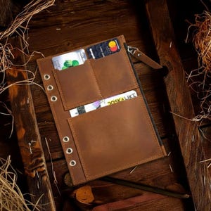 May include: A brown leather wallet with multiple card slots and a zippered compartment. The wallet is open, revealing credit cards and a business card. Metal eyelets line the left side. The wallet is on a wooden surface.