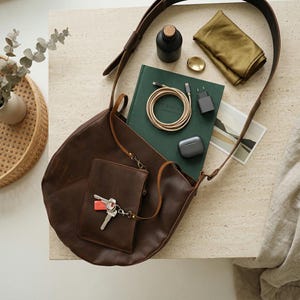 May include: A dark brown leather shoulder bag with an attached small pouch featuring a set of keys. The bag is surrounded by a dark green notebook, a coiled charging cable, a black wall adapter, a small black bottle, and a folded olive cloth.