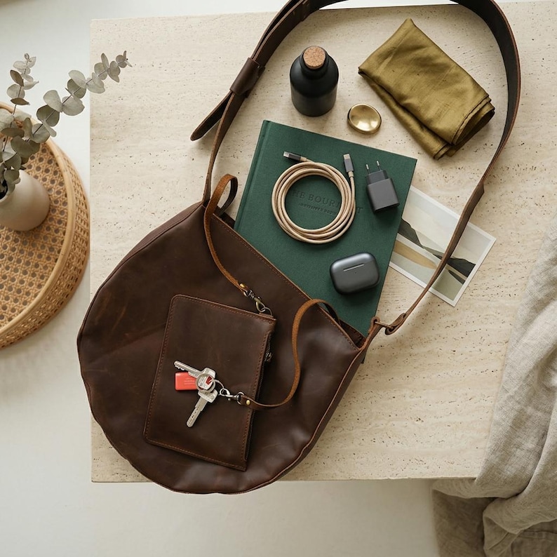 May include: A dark brown leather shoulder bag with an attached small pouch featuring a set of keys. The bag is surrounded by a dark green notebook, a coiled charging cable, a black wall adapter, a small black bottle, and a folded olive cloth.