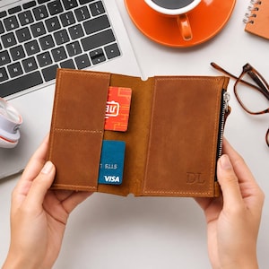 May include: A brown leather wallet featuring two vertical card slots on the left side, one containing a red card and one containing a blue Visa card, and a zippered compartment on the right side with the embossed initials DL on the front.