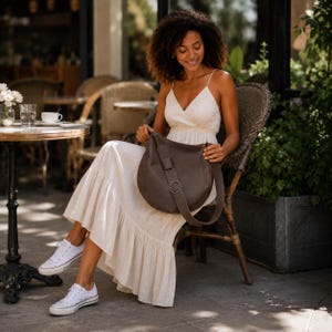 May include: A dark brown leather hobo bag with a wide shoulder strap and a circular metal ring detail. The bag features a smooth, rounded silhouette and is worn across the body. It is paired with a sleeveless cream-colored maxi dress and white sneakers.