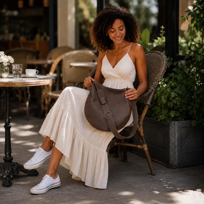 May include: A dark brown leather hobo bag with a wide shoulder strap and a circular metal ring detail. The bag features a smooth, rounded silhouette and is worn across the body. It is paired with a sleeveless cream-colored maxi dress and white sneakers.