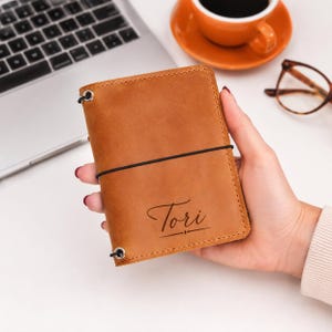 Puede incluir: Una funda de cuaderno de cuero color canela con un cierre de banda elástica negra y el nombre Tori grabado en letra cursiva en la parte frontal. La funda incluye ojales metálicos en las esquinas para asegurar el cordón elástico vertical.