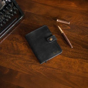 Puede incluir: Una cartera de cuero negro con cierre de bot&oacute;n a presi&oacute;n y el nombre "Kate" grabado en letras doradas. Un bol&iacute;grafo de madera con detalles dorados y un capuch&oacute;n est&aacute;n junto a la cartera sobre una superficie de madera. Una m&aacute;quina de escribir vintage est&aacute; en el fondo.