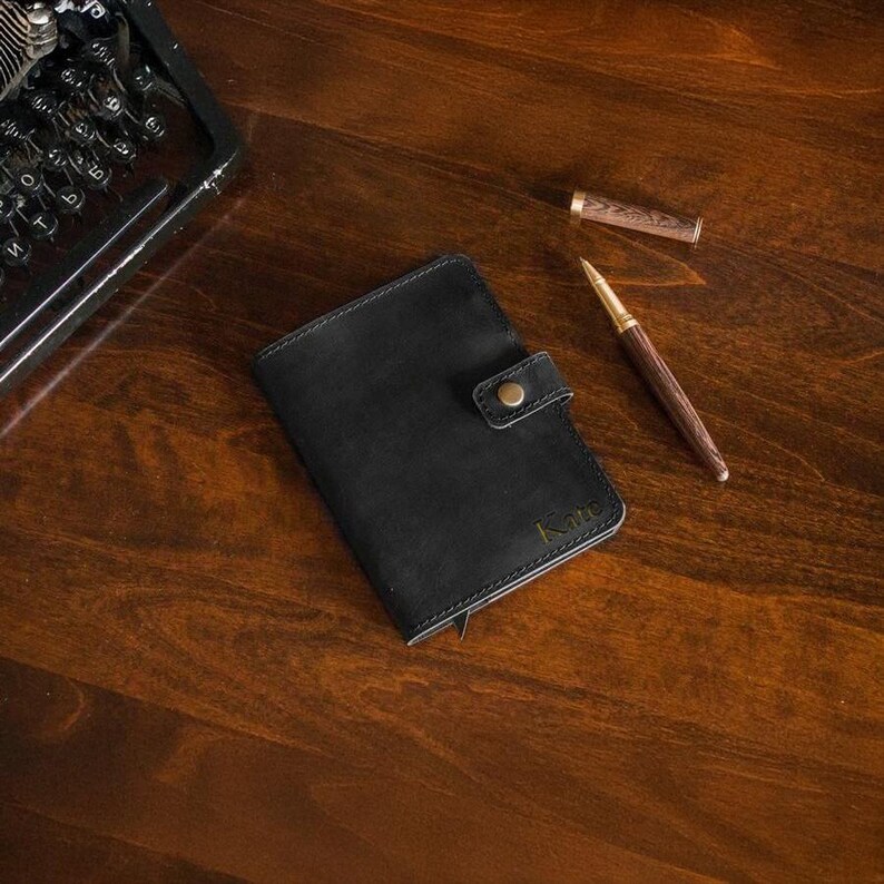 Puede incluir: Una cartera de cuero negro con cierre de bot&oacute;n a presi&oacute;n y el nombre "Kate" grabado en letras doradas. Un bol&iacute;grafo de madera con detalles dorados y un capuch&oacute;n est&aacute;n junto a la cartera sobre una superficie de madera. Una m&aacute;quina de escribir vintage est&aacute; en el fondo.