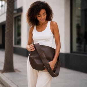 May include: A dark brown hobo bag with a wide shoulder strap featuring a large circular metal buckle. The bag has a smooth, matte finish and a slouchy, rounded shape. It is worn over the shoulder, resting against the torso, showcasing the hardware.