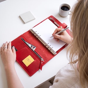 Puede incluir: Una agenda de cuero rojo brillante con un mecanismo de encuadernación de metal de seis anillas, que incluye un bolsillo interior con cremallera, una ranura para tarjetas con una tarjeta amarilla, un portalápices con un bolígrafo plateado y hojas.