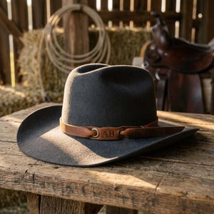 Könnte beinhalten: Ein anthrazitfarbener Cowboyhut aus Filz mit einem hellbraunen Lederhutband, das mit messingfarbenen Metallösen und den mittig eingeprägten Buchstaben AB versehen ist. Der Hut hat eine strukturierte Krone und eine breite, geschwungene Krempe.