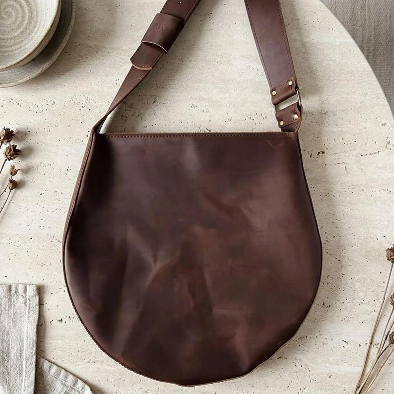 May include: A dark brown leather shoulder bag with a smooth, matte finish and a rounded, semi-circular silhouette. The bag features a wide, adjustable leather strap attached with metallic hardware and visible stitching along the top edge.