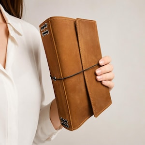 May include: A rectangular tan leather journal featuring a smooth matte finish, secured by a thin black elastic cord wrapped horizontally around the cover. The spine area includes metal eyelets with black cord lacing at the top and bottom edges.