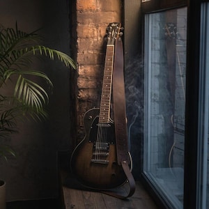 K&ouml;nnte beinhalten: Eine Cort E-Gitarre mit dunkelbraunem Sunburst-Finish und schwarzem Schlagbrett, ausgestattet mit einem langen, schlichten braunen Ledergurt, der an Kopfplatte und Korpus befestigt ist und aufrecht an einer strukturierten Ziegelwand neben einem Fensterrahmen steht.