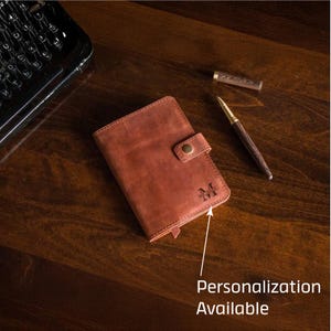 Puede incluir: Un diario de cuero marr&oacute;n con cierre de bot&oacute;n, personalizado con la letra "M". Un bol&iacute;grafo y una tapa de bol&iacute;grafo est&aacute;n junto al diario. El texto "Personalizaci&oacute;n Disponible" est&aacute; en la imagen. El diario est&aacute; sobre una superficie de madera.