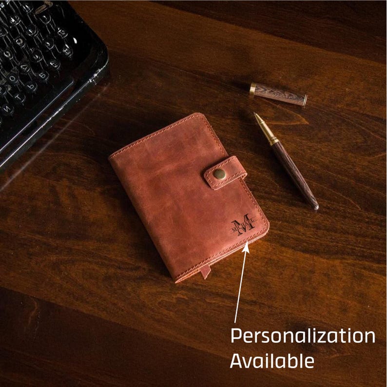 Puede incluir: Un diario de cuero marr&oacute;n con cierre de bot&oacute;n, personalizado con la letra "M". Un bol&iacute;grafo y una tapa de bol&iacute;grafo est&aacute;n junto al diario. El texto "Personalizaci&oacute;n Disponible" est&aacute; en la imagen. El diario est&aacute; sobre una superficie de madera.