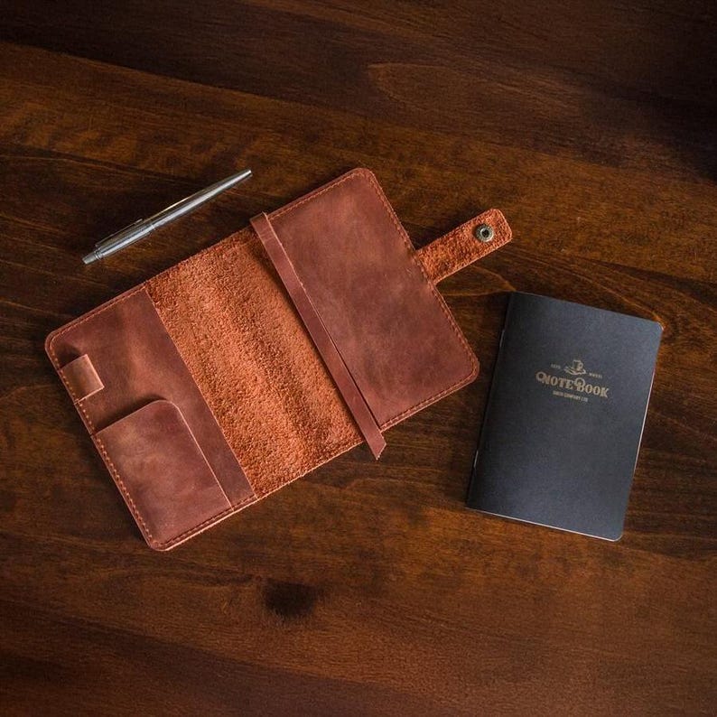 Puede incluir: Una funda de cuero marr&oacute;n para cuaderno, un bol&iacute;grafo plateado y un cuaderno negro sobre una superficie de madera. La funda est&aacute; abierta, revelando un bolsillo y una correa. El cuaderno negro tiene el texto "NOTEBOOK" en dorado.